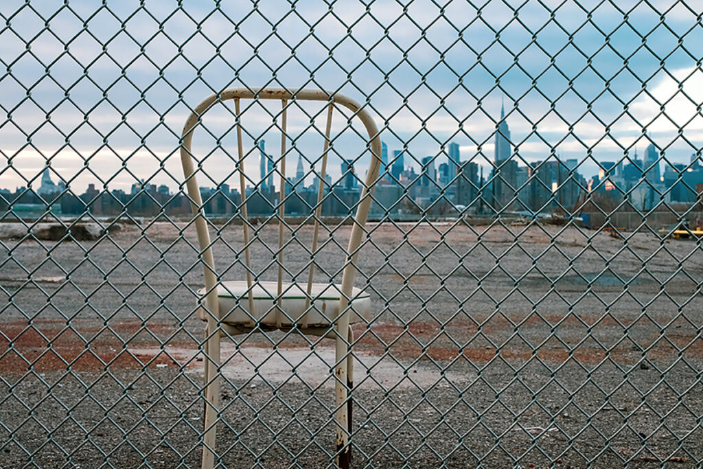 A chainlink fence - through which you see a metal hooped back white chair on a derelict empty lot, with the skyline of New York seen from Brooklyn/Queens