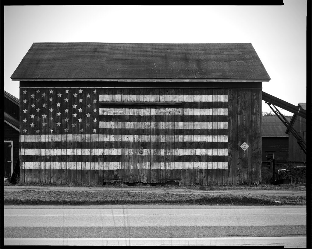 Black and white photograph of the side of a barn,  on which an American flag has been painted