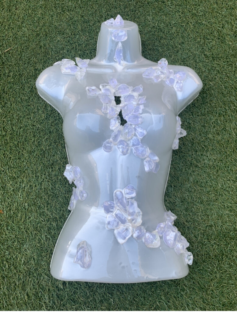 photograph of a opaque plastic female body form  lying on  the grass.  The torso form is scattered with large cyrstal like shapes.