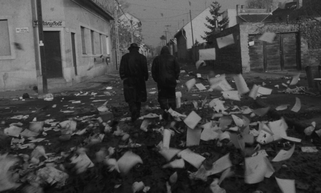 Black and white film still of two men in coats, one with a hat, walking away from the viewer, down a town street, strewn with lots of paper