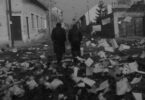 Black and white film still of two men in coats, one with a hat, walking away from the viewer, down a town street, strewn with lots of paper.