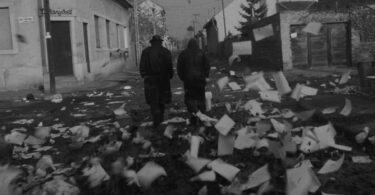 Black and white film still of two men in coats, one with a hat, walking away from the viewer, down a town street, strewn with lots of paper.
