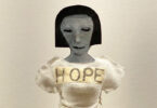 Image of a doll with a short haircut. She is dressed in a white dress with the word HOPE written across her chest