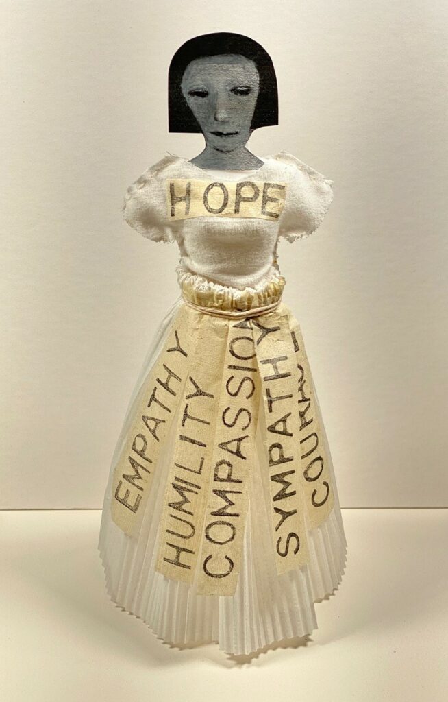 Image of a doll with a charcoal black head of hair and face.  She is dressed in a creamy white dress with the word HOPE written across her chest and the words 
Empathy, Humility, Compassion, Sympathy and Courage written on strips hanging from her waist