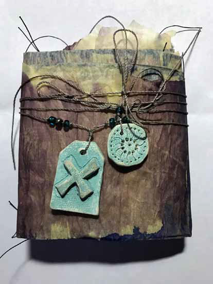 A book like assemblage wrapped in altered issue paper, tied in twine with turquoise beads and two blue objects made of clay.