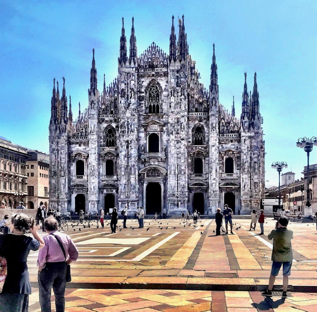 Photograph of Milan Cathedral and Piazza del Duomo.  The magnificent cathedral is in the background.  A few people are standing around, there are more pigeons on the ground than people.  There are a few poeple with masks.  