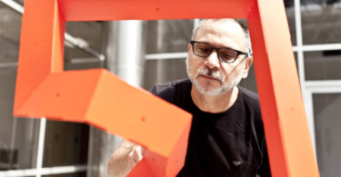 Man wearing glasses looking at an abstract sculpture with windows behind him.