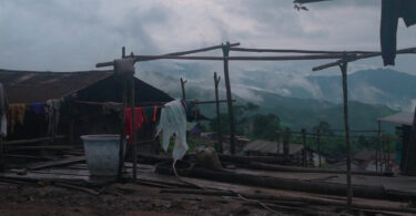 Wooden poles creating a structure with clothes hanging on it. Clouds and mountains are seen in the background.