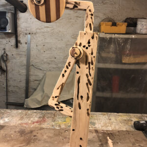 photograph of a wooden sculpture in a workshop. the piece has a circular head and an arm with a long body