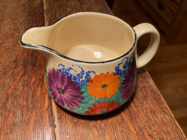 round squat jug with a handle and bright daisy like flowers on a green grass packground - jug is on a wooden floor.