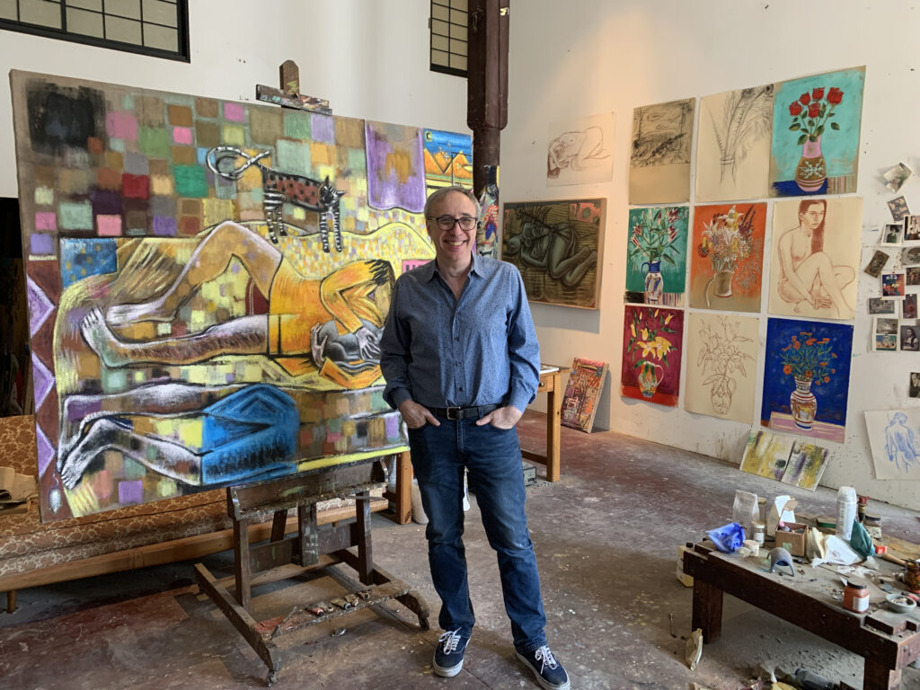 photograph of Murk Kurdziel in a blue shirt and blue jeans in his studio.  Paintings on an easel behind his right shoulder and drawings on the wall behind his left shoulder.  Ta low table to his side is covered with paints, cloths, brushes, jars and bottles.
