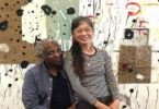 Photo of Carl Heyward sitting and Akiko Suzuki standing and leaning against Carl with mixed media art on the wall behind them