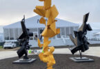 outdoor photo of three metal sculptures, yellow in the center and two black sculptures either side. A large tented building stands behind