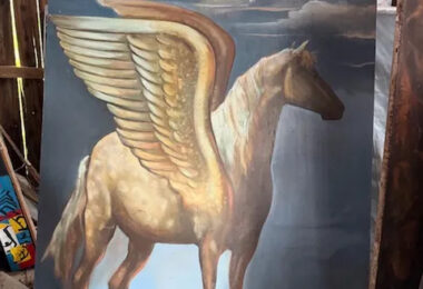 Oil painting of pegasus standing on a field with wings lifted up with a blue sky and clouds in the background. The painting is in a wooden barn.